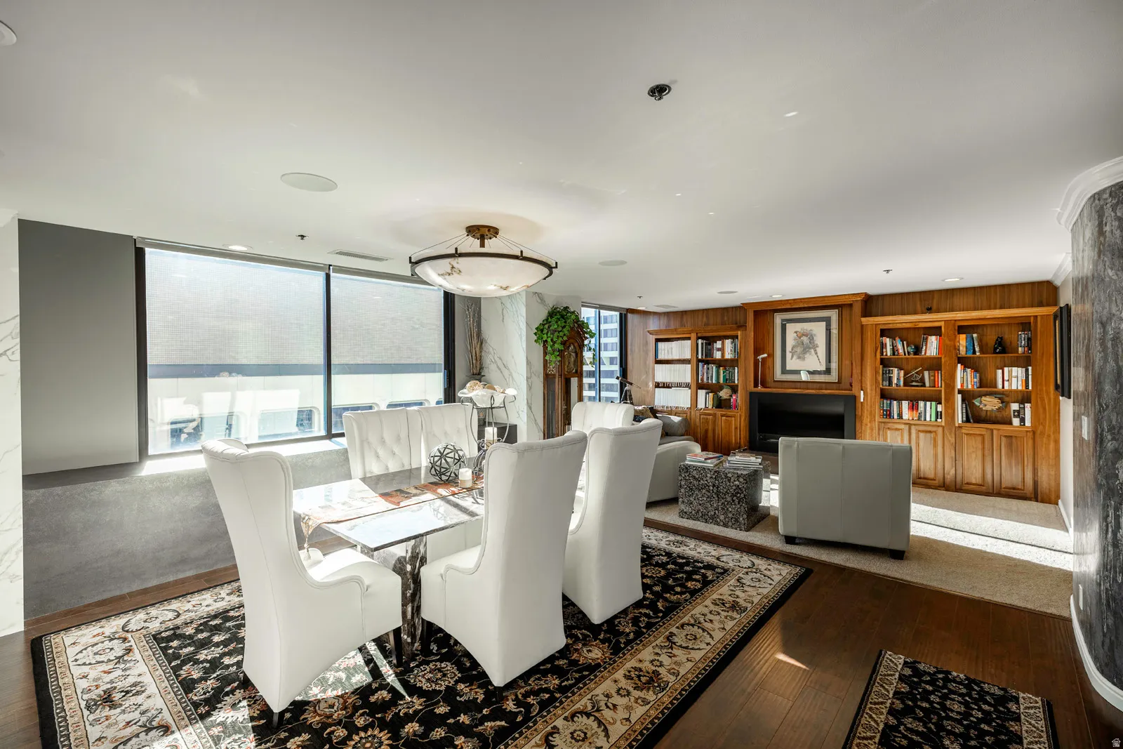 Open-concept living and dining area featuring wood-finish flooring, large windows with roller shades, a built-in fireplace with a dark surround, custom wood built-in shelving, and recessed lighting