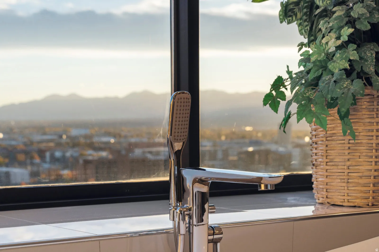 Modern chrome freestanding tub filler with integrated handheld shower, positioned against a large window featuring city and distant mountain scenery