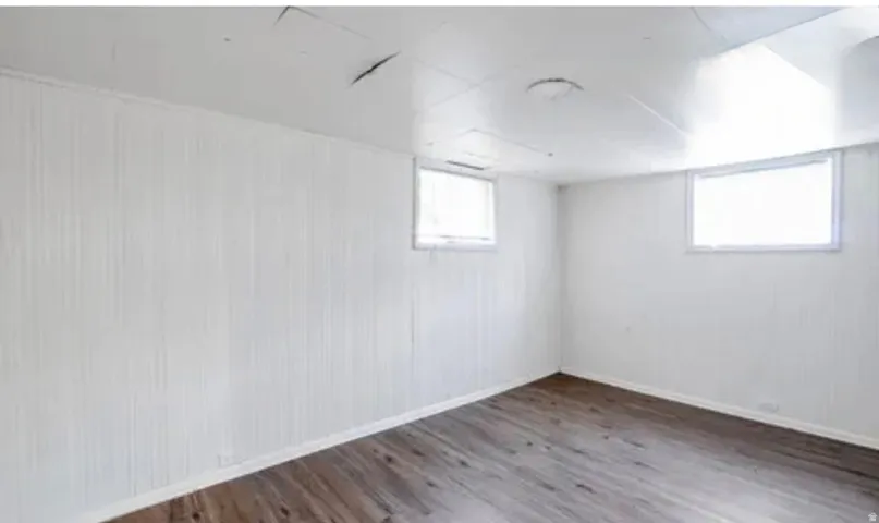Spacious room featuring wood-finish flooring, white paneled walls, and two windows providing natural illumination