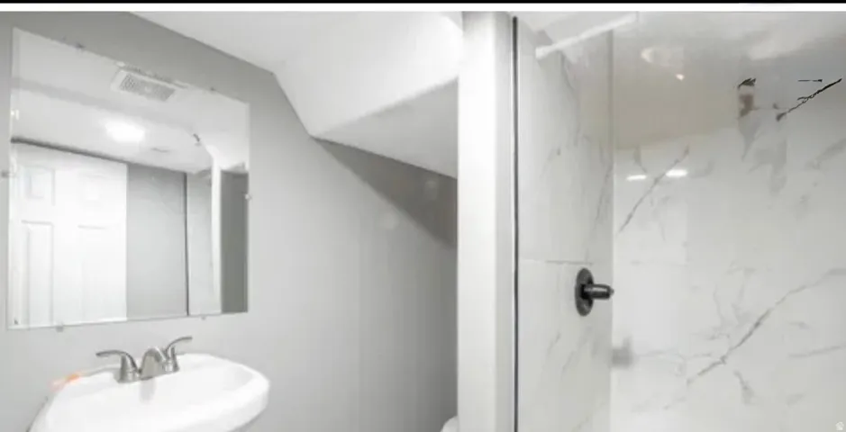 Bathroom featuring a pedestal sink with a chrome faucet, wall-mounted mirror, and a shower with white veined tile surround and a matte black shower fixture