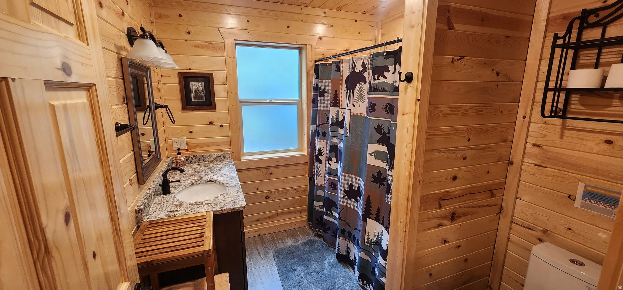 Bathroom featuring natural wood-finish plank walls and ceiling, a single vanity with a solid surface countertop and undermount sink, a bronze-finish faucet, and a shower area with a frosted window