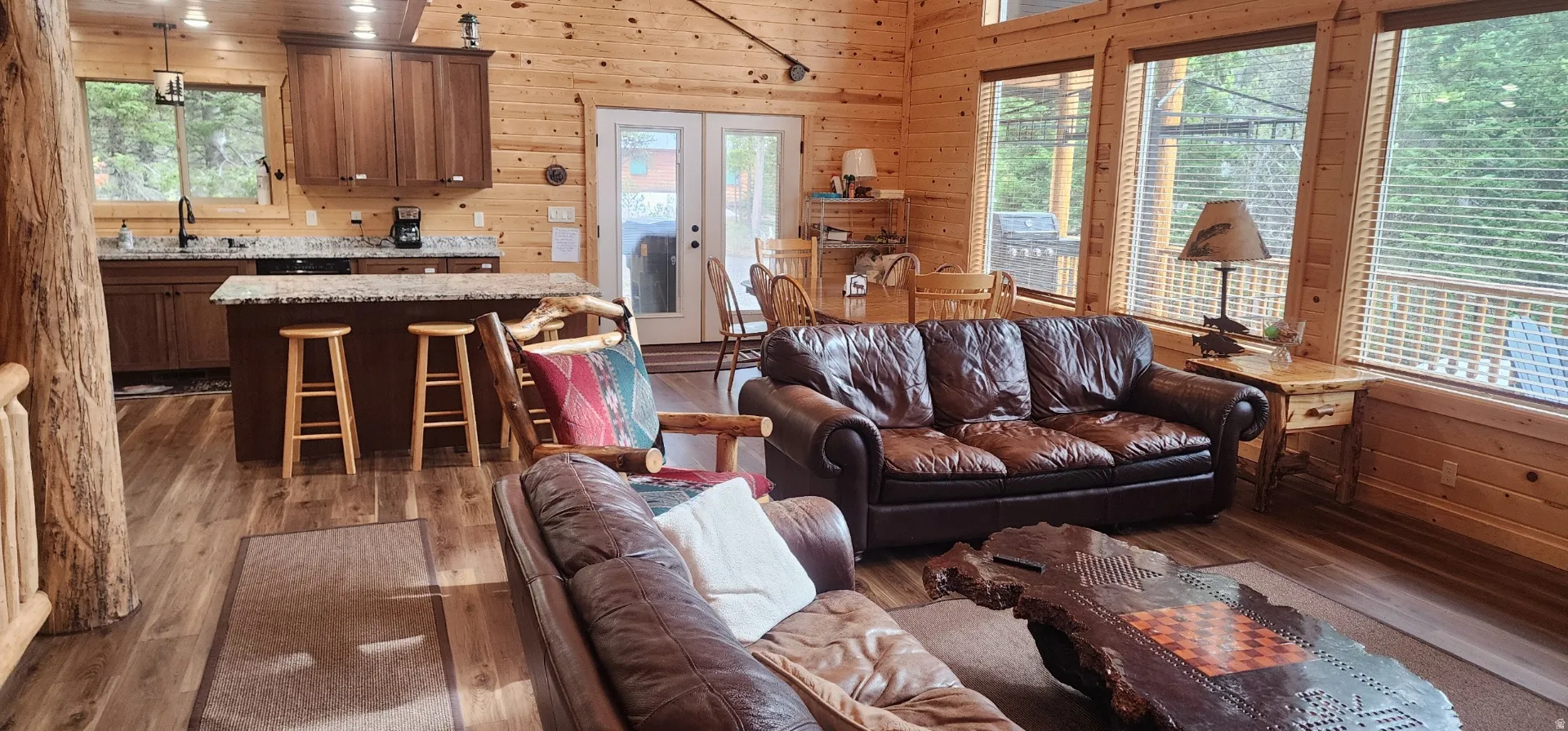Open-concept living space featuring wood-finish flooring, natural wood paneling, a kitchen island with seating, a full kitchen with dark cabinetry, and multiple large windows