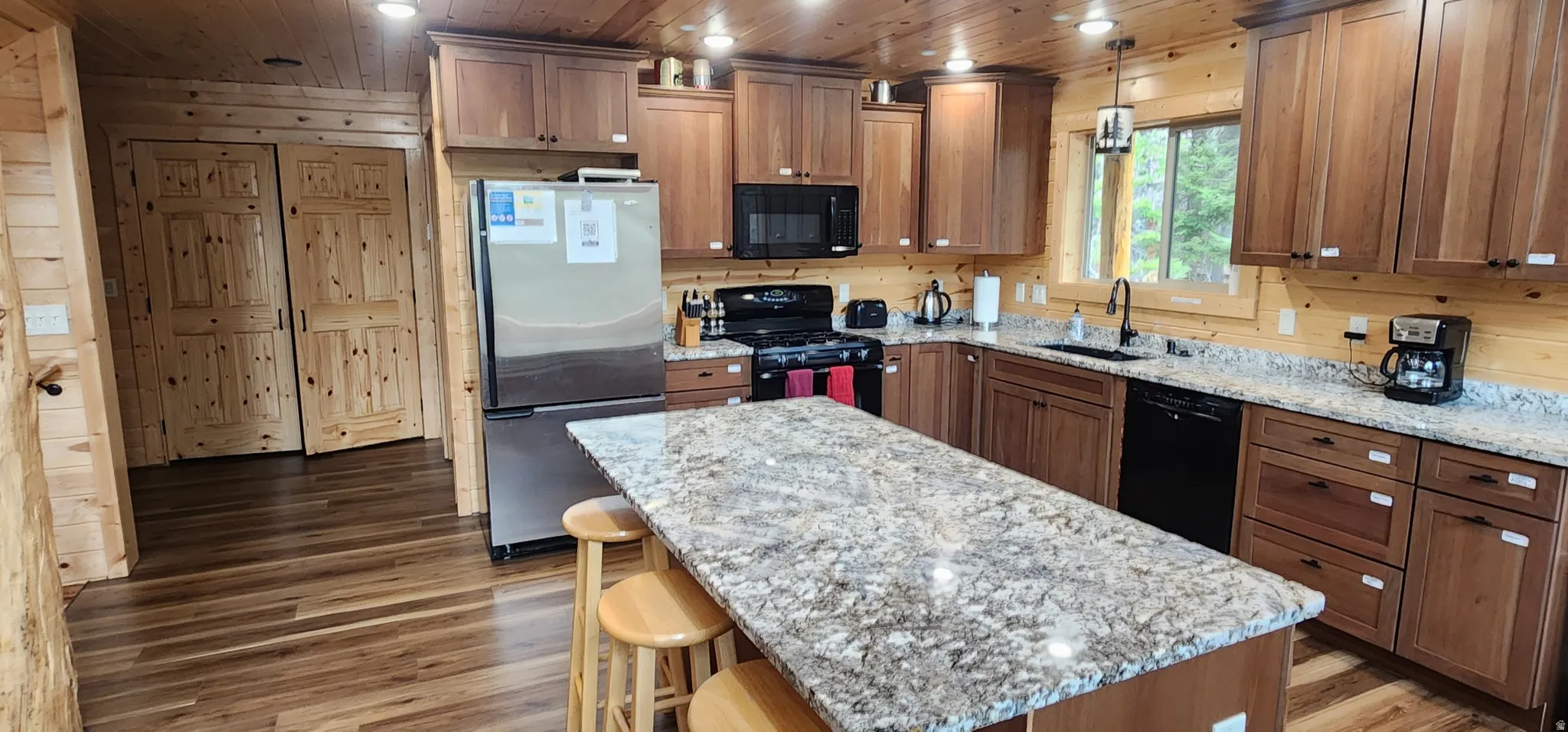 Kitchen featuring a substantial island with a light-toned patterned countertop, wood-finish cabinetry, stainless steel refrigerator, black range, and black dishwasher