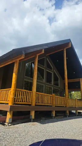 Modern cabin-style exterior featuring a dark gray roofline, a full-length wooden deck with natural log railings, and a prominent A-frame window design