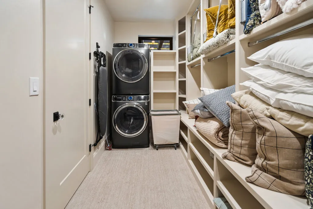 Dedicated laundry area featuring stacked black appliances, integrated shelving, and light-colored carpet flooring