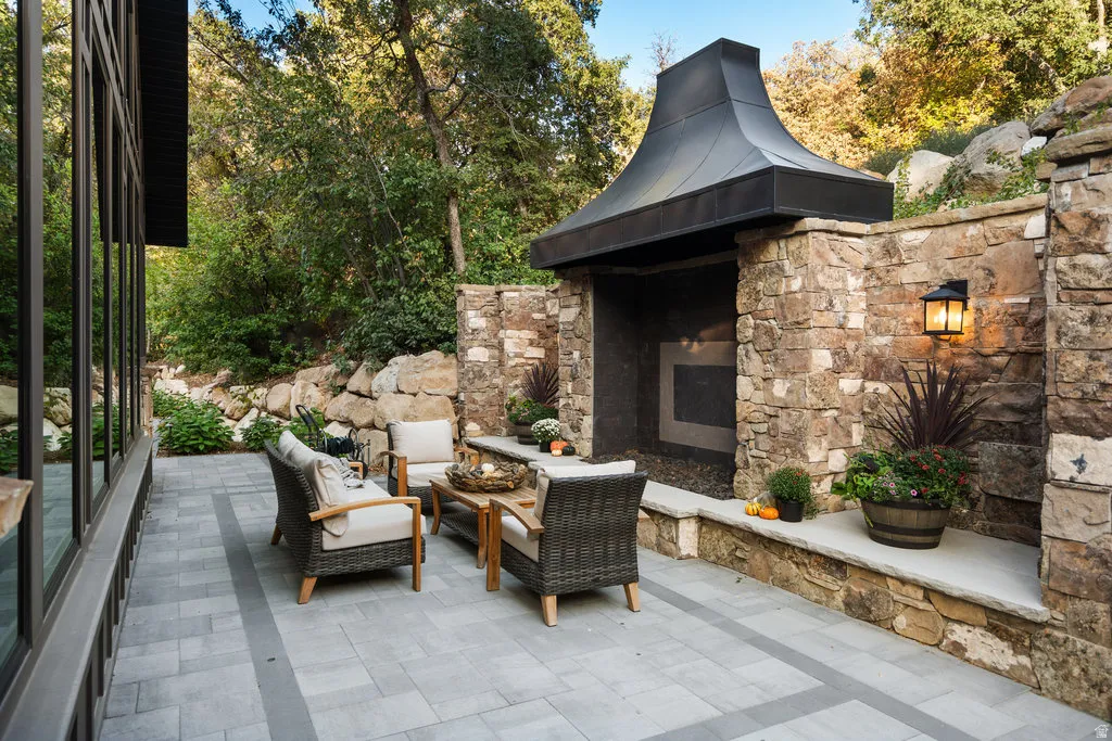 Expansive outdoor patio featuring paver flooring, a built-in stone fireplace with a metal hood, and a stone retaining wall
