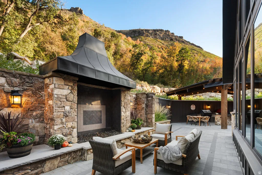 Expansive hardscaped patio featuring a stone fireplace with a metal hood, a covered outdoor kitchen area, and large format windows