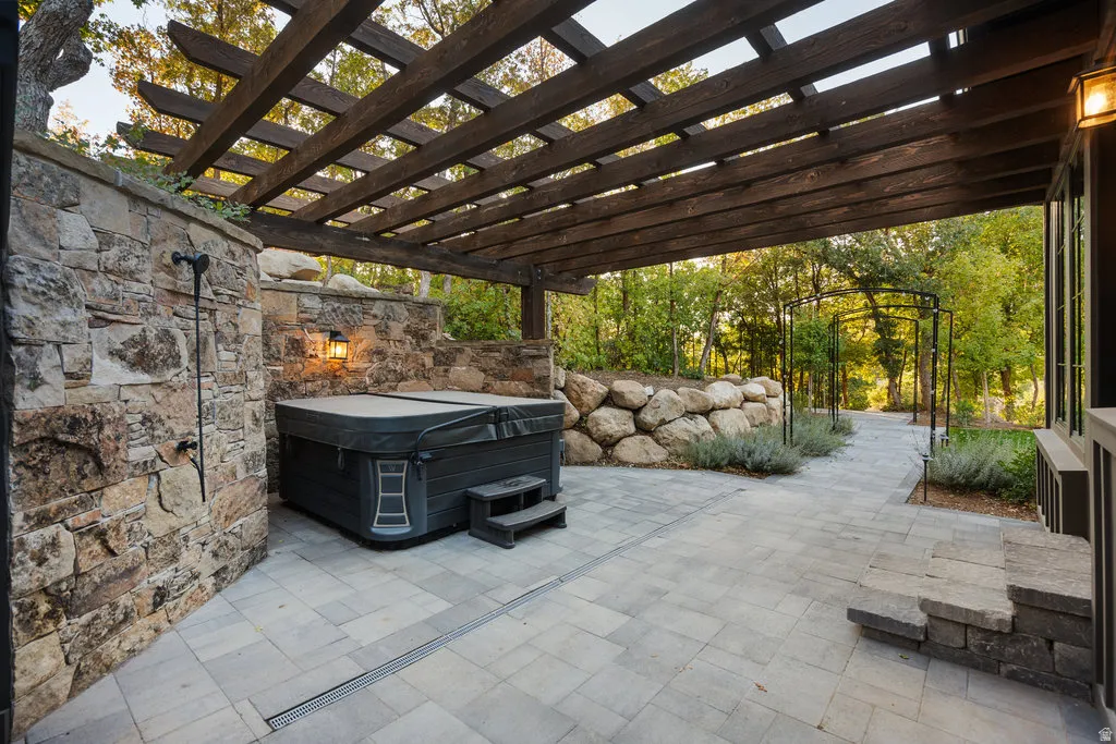 Expansive stone paver patio featuring a hot tub and a dark wood pergola