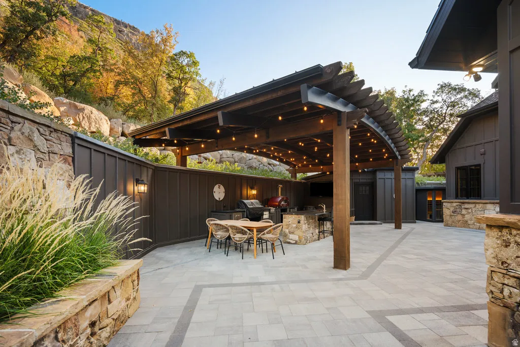 Expansive paved patio featuring a curved wood pergola with string lighting, an outdoor kitchen with a built-in grill and stone-clad bar, and a stone retaining wall with mature plantings