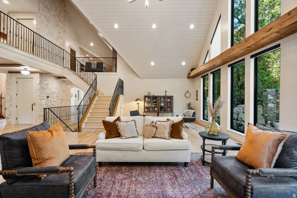 Vaulted ceiling living area featuring a stone accent wall, wood-finish flooring, a grand staircase with wrought iron railings, and extensive windows with wood beam accents