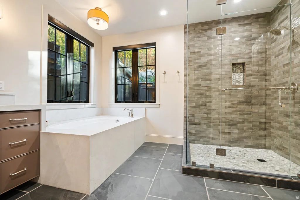 Spacious bathroom featuring a frameless glass shower enclosure, a built-in soaking tub, dark slate tile flooring, a built-in vanity with drawer storage, and two large windows with dark trim