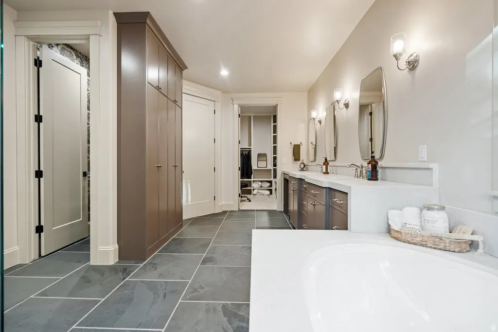 Spacious bathroom featuring large format stone tile flooring, a built-in soaking tub, and a double vanity with undermount sinks and wall-mounted sconces