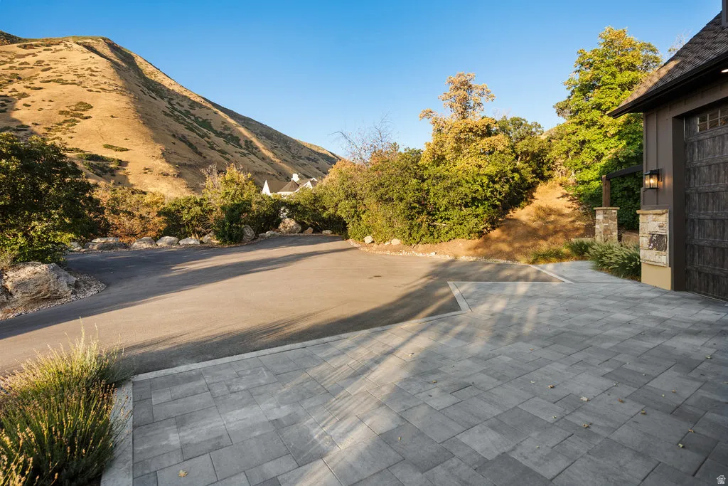 Paver driveway with an integrated asphalt approach, bordered by mature landscaping and a stone-clad pillar with an exterior sconce
