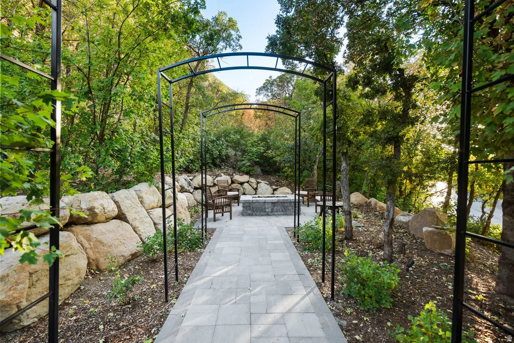 Outdoor patio featuring a stone-paved walkway, multiple metal arbors, natural rock retaining walls, a built-in fire pit, and extensive natural landscaping