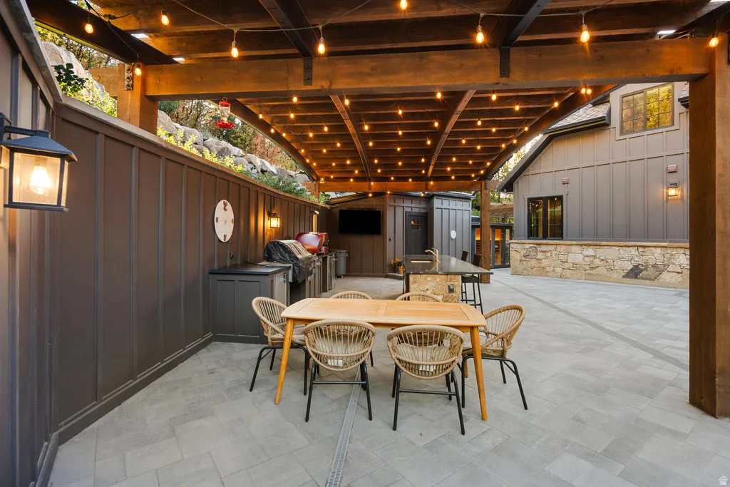 Expansive outdoor patio featuring a wooden pergola with string lighting, board and batten wall, paved flooring, and a built-in outdoor kitchen with a grill and sink