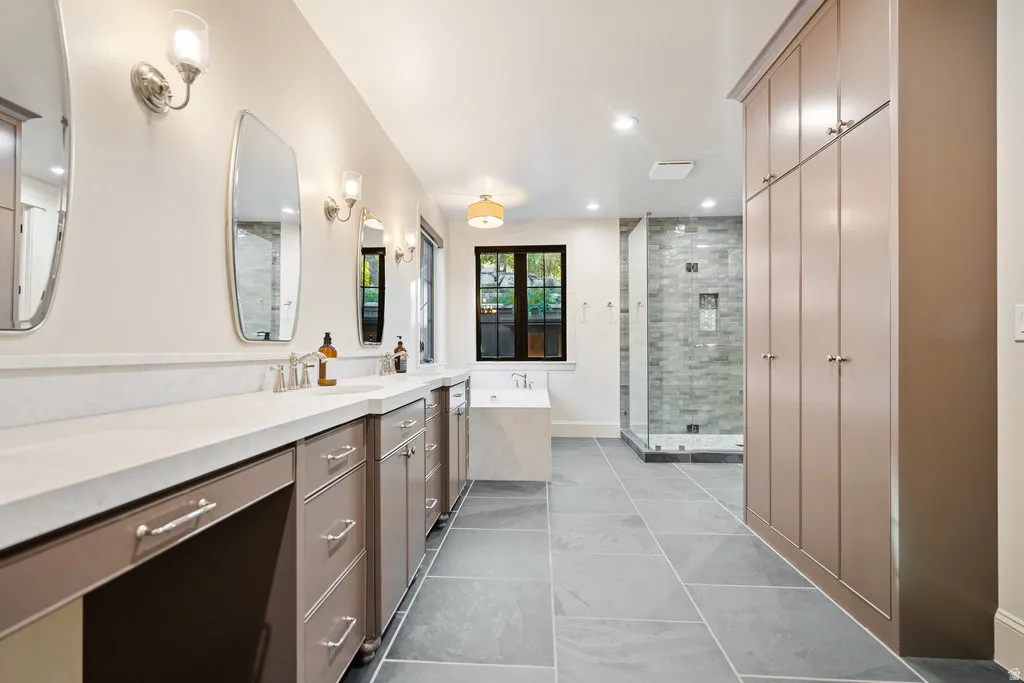 Spacious bath featuring a dual vanity with light countertops, extensive built-in cabinetry, an enclosed shower with a tiled surround, a freestanding soaking tub, and large format floor tiling