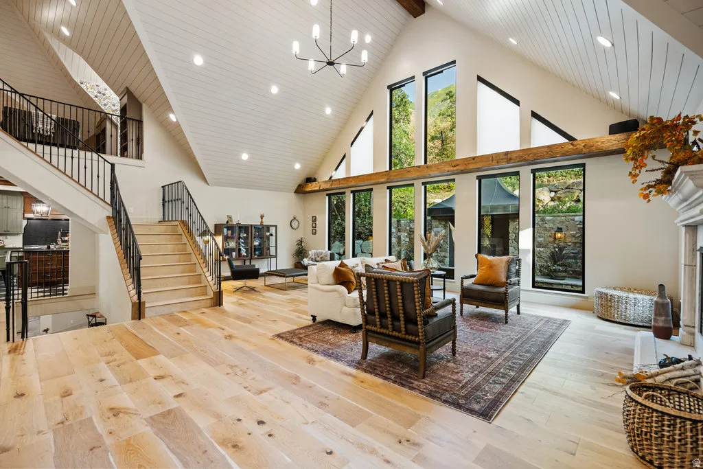 Spacious living area featuring high vaulted ceilings with exposed wood beams, light wood-finish flooring, a grand staircase with wrought iron balusters, a stone fireplace with a substantial mantel, and multiple large windows with black framing