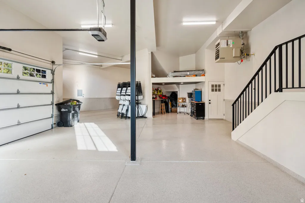 MAIN pacious garage featuring a light-colored epoxy floor, overhead lighting, a white panel garage door with windows, a white interior door, and a black metal handrail ascending a staircase
