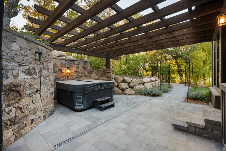 Expansive stone paver patio featuring a hot tub and a dark wood pergola