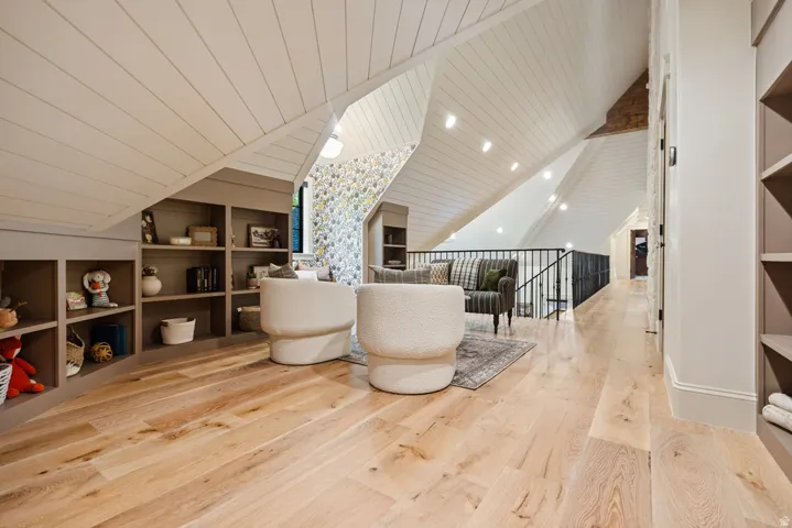 Spacious loft area featuring light wood-finish flooring, built-in shelving, vaulted shiplap ceilings with recessed lighting, and a black metal railing