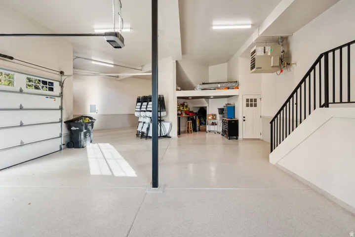 MAIN pacious garage featuring a light-colored epoxy floor, overhead lighting, a white panel garage door with windows, a white interior door, and a black metal handrail ascending a staircase