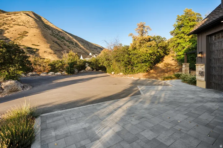 Paver driveway with an integrated asphalt approach, bordered by mature landscaping and a stone-clad pillar with an exterior sconce