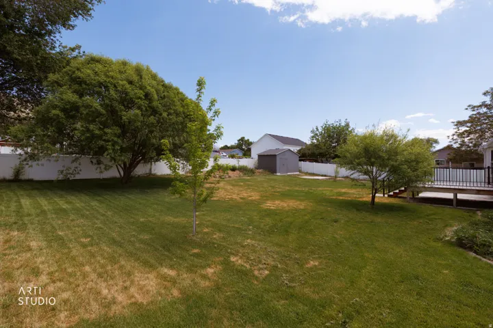 Expansive backyard featuring a white privacy fence, mature trees, a wooden deck with black railings, and a detached storage shed