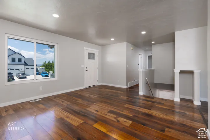 Spacious open-concept room featuring wide-plank wood-finish flooring, recessed ceiling lighting, a white entry door with a decorative glass insert, and a large window providing ample natural light