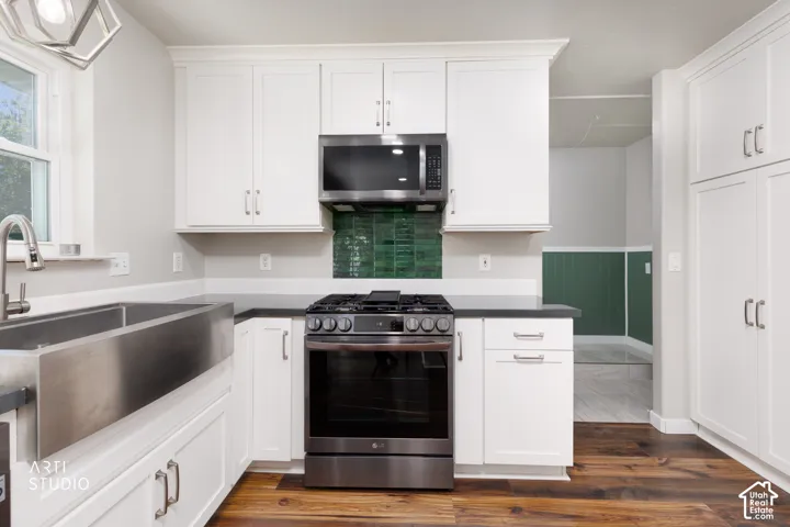 Kitchen featuring white shaker cabinetry, dark countertops, stainless steel appliances, a farmhouse sink, and wood-finish flooring