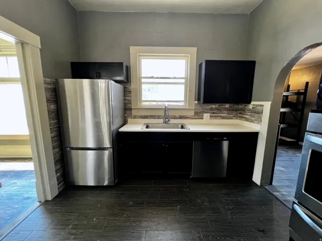Kitchen featuring a stainless steel refrigerator, dark wood-finish flooring, black cabinetry, light countertops, and a tiled backsplash