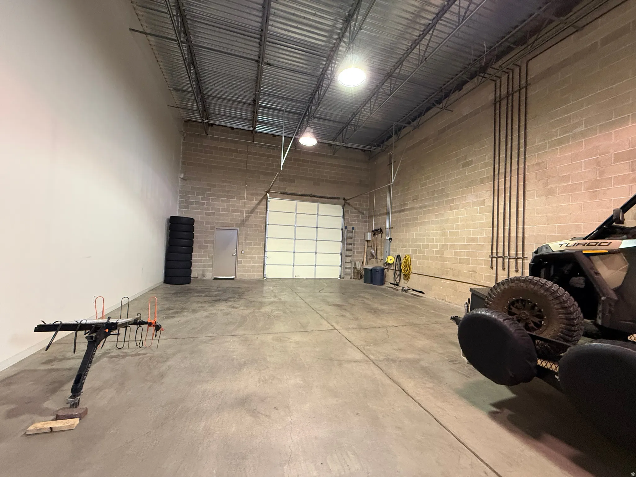 Spacious interior featuring a large roll-up garage door, concrete flooring, exposed overhead piping, block walls, and high ceilings with industrial lighting
