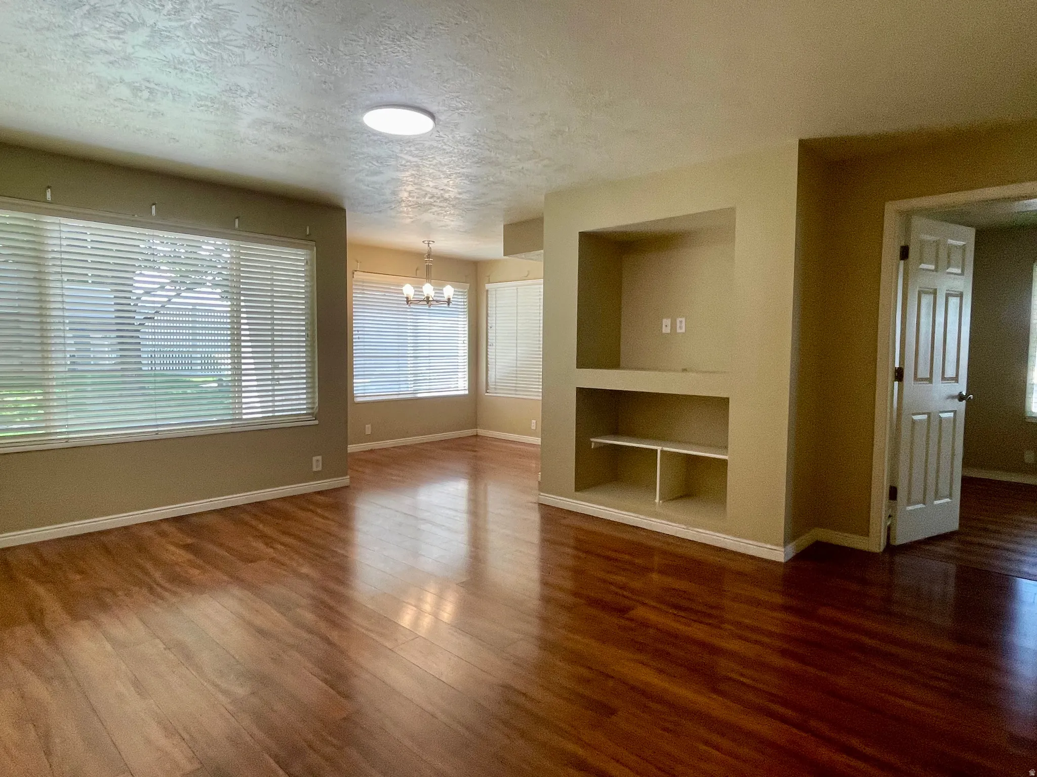 Spacious living area featuring rich wood-finish flooring, multiple windows with horizontal blinds, and a built-in media niche with shelving
