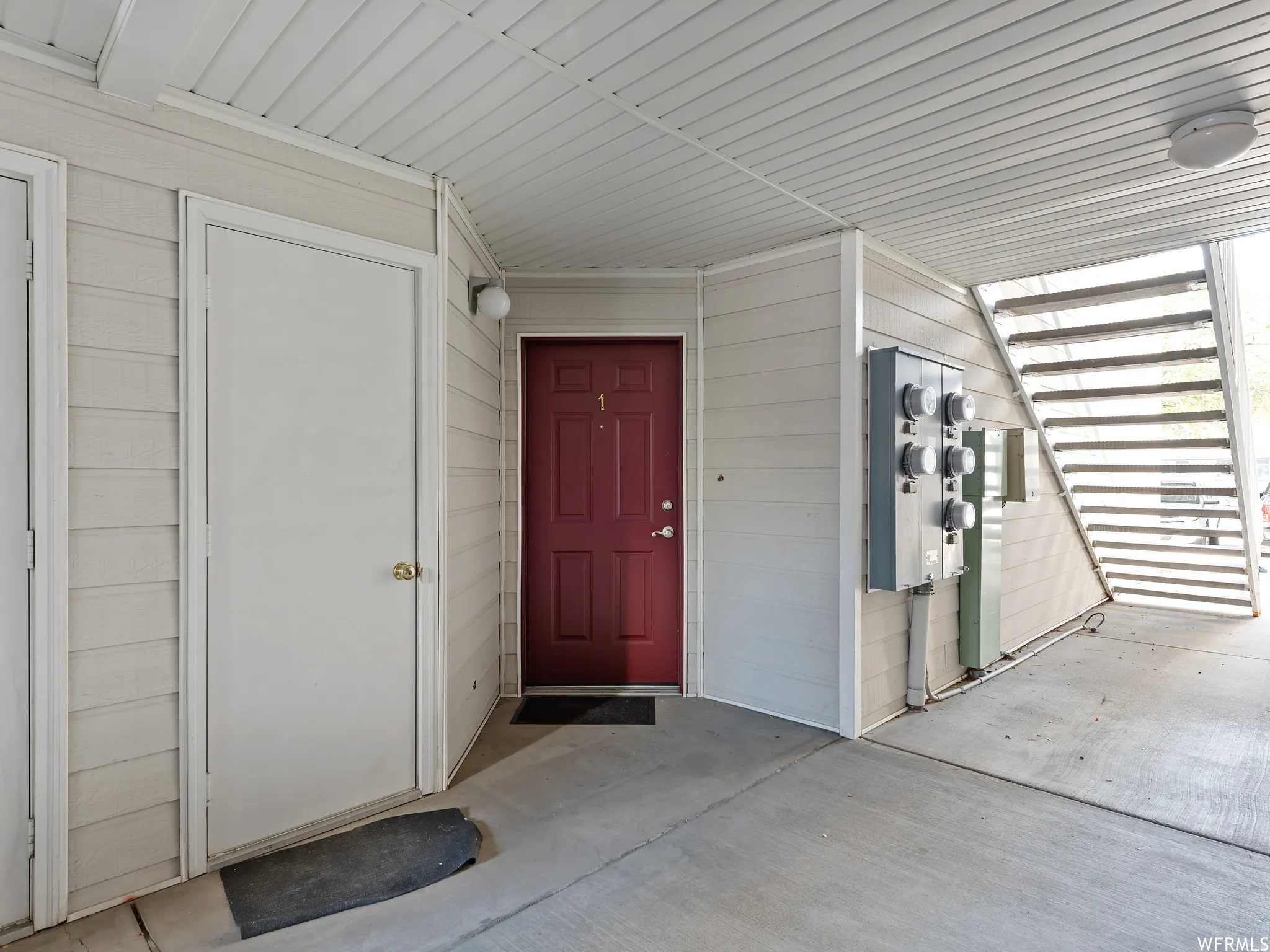 Exterior entryway featuring a red door with numerical designation, light-colored siding, concrete flooring, and an overhead lighting fixture