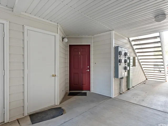 Exterior entryway featuring a red door with numerical designation, light-colored siding, concrete flooring, and an overhead lighting fixture