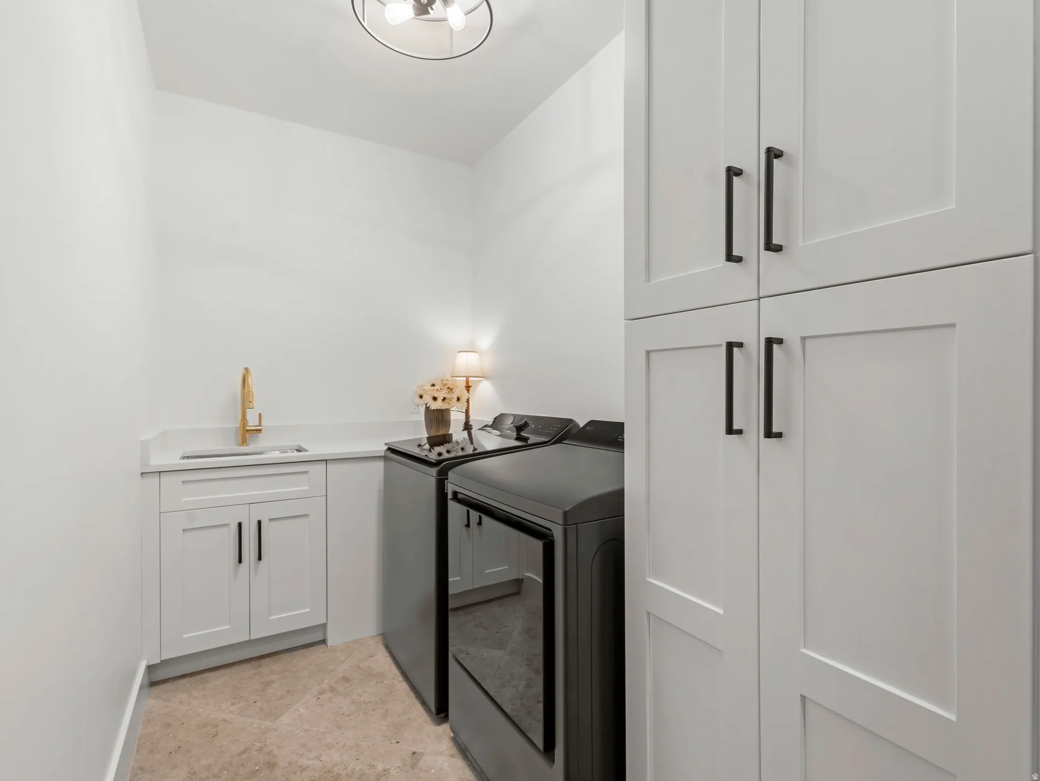 Dedicated laundry area featuring a utility sink with a gold-finish faucet, white cabinetry with black hardware, white countertops, and light-toned floor tiling