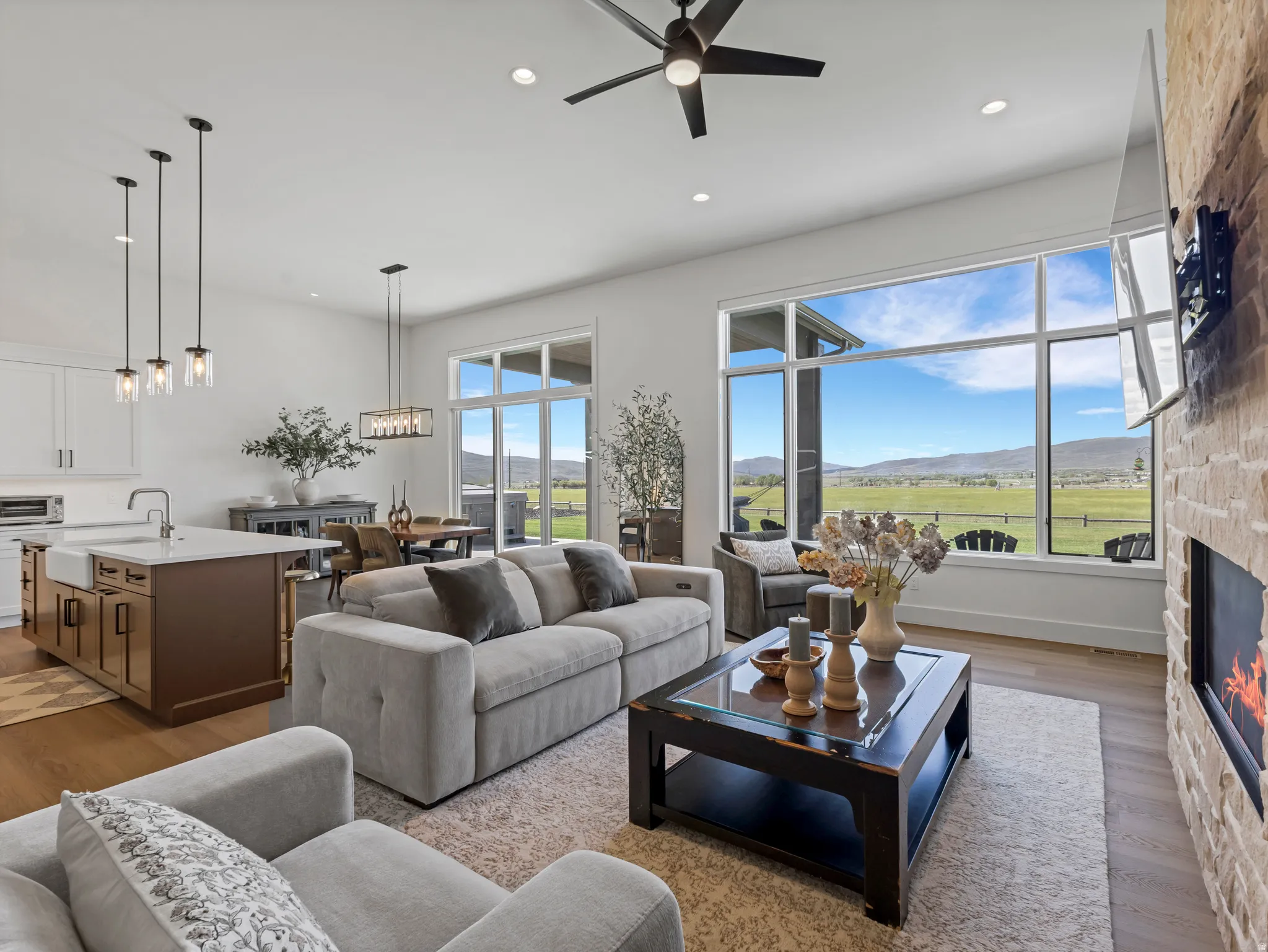 Open-concept interior featuring light wood-finish flooring, a stone-clad fireplace, and expansive windows