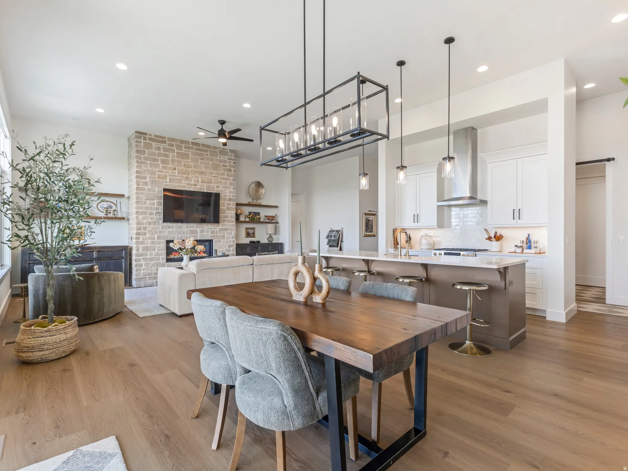 Open-concept living space featuring wood-finish flooring, a stone-clad fireplace with built-in shelving, a kitchen island with seating, pendant lighting, and recessed lighting