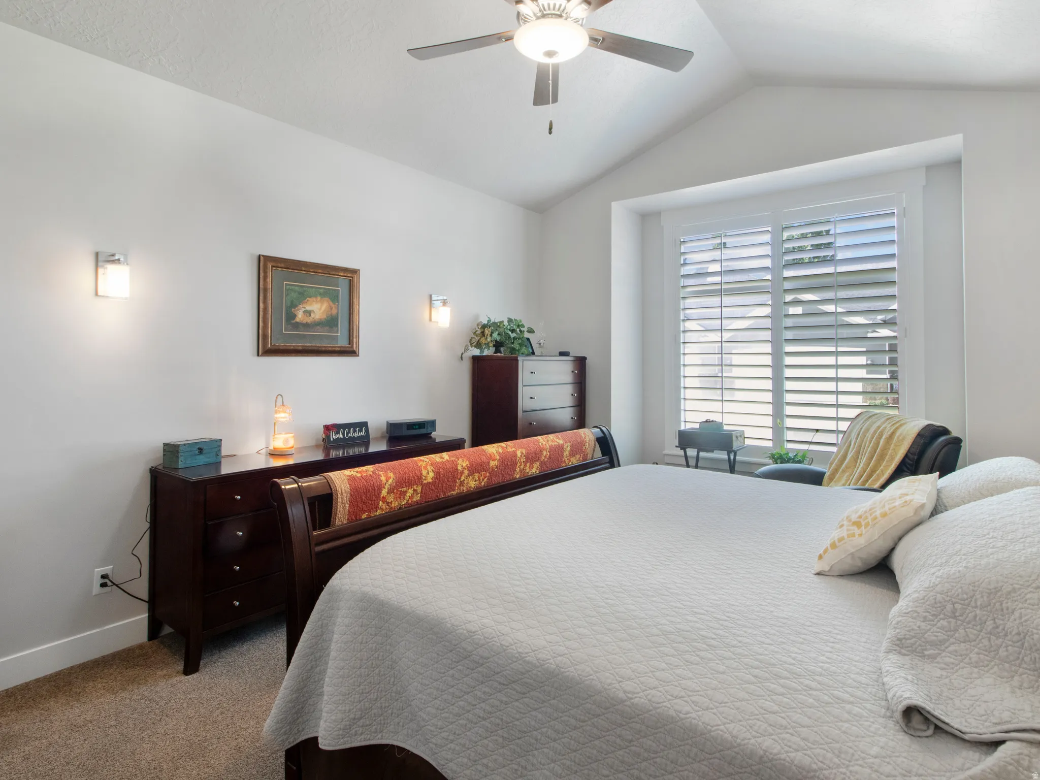 Vaulted ceiling bedroom with a ceiling fan, sconce lighting, and a window featuring white plantation shutters