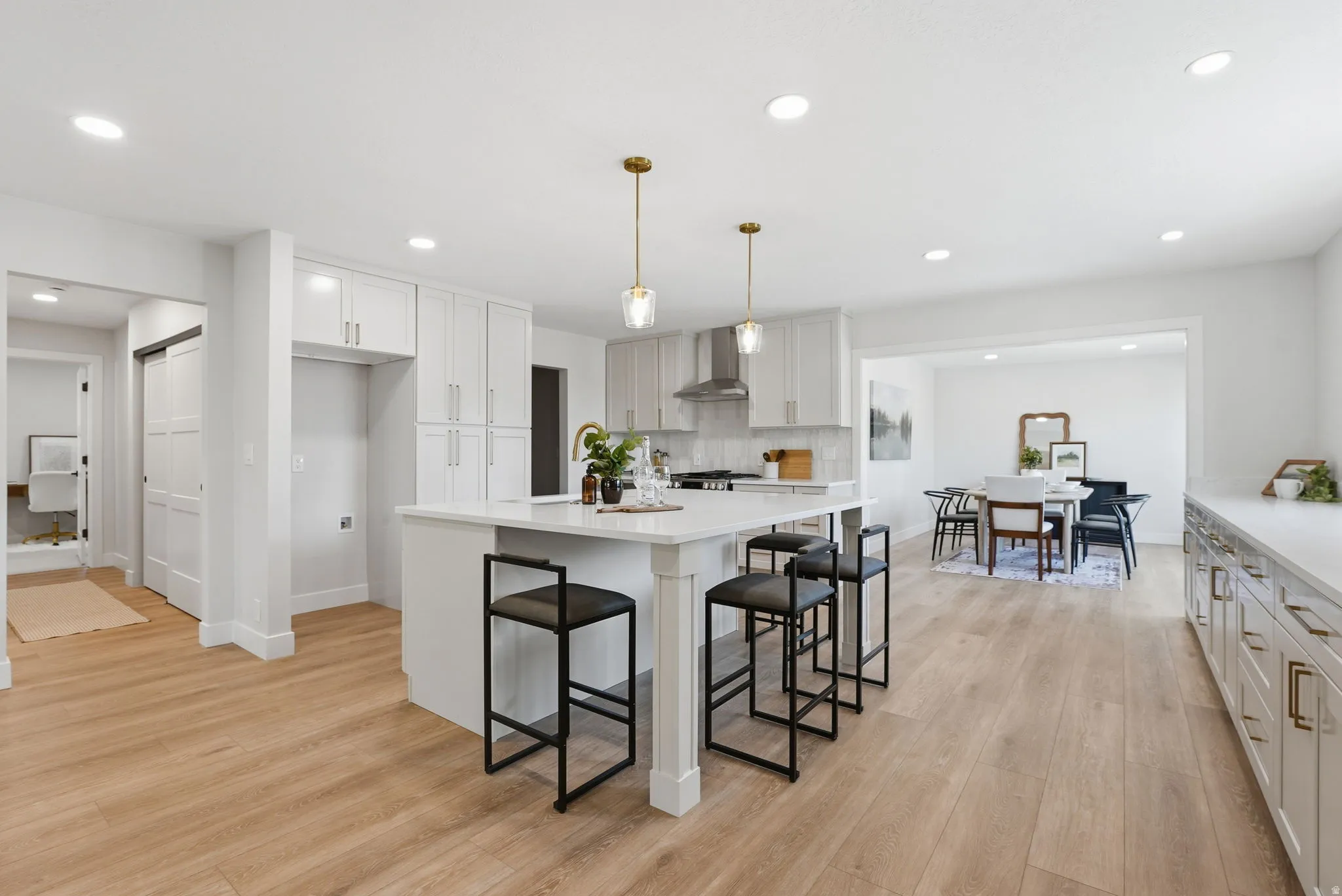 Open-concept kitchen featuring an expansive island with seating, white shaker cabinetry, gold-finish hardware, stainless steel range hood, and wood-finish flooring