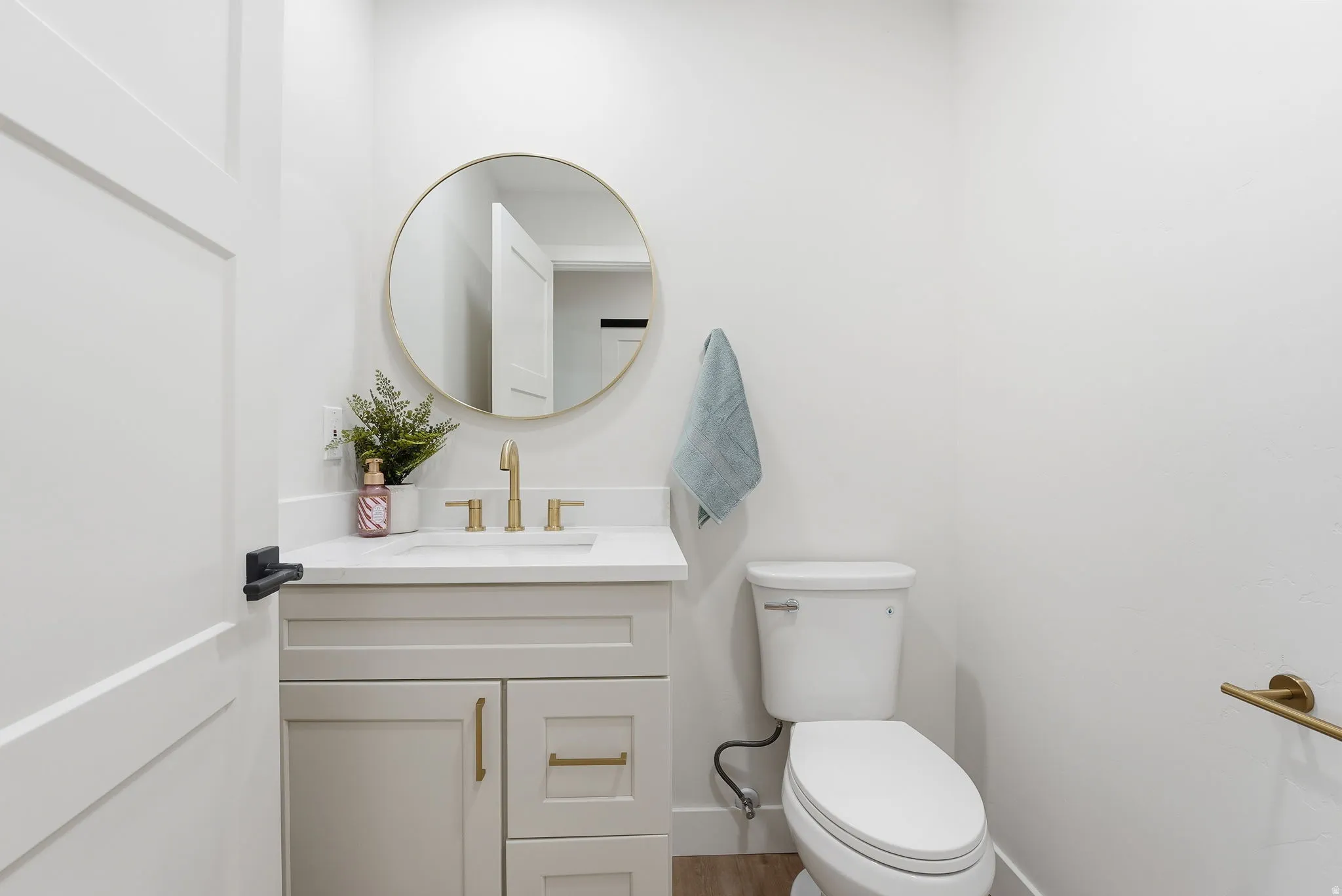 Contemporary powder room featuring a single-panel door with black hardware, a light gray vanity with gold-tone pulls, a white countertop, a circular gold-framed mirror, and a modern toilet