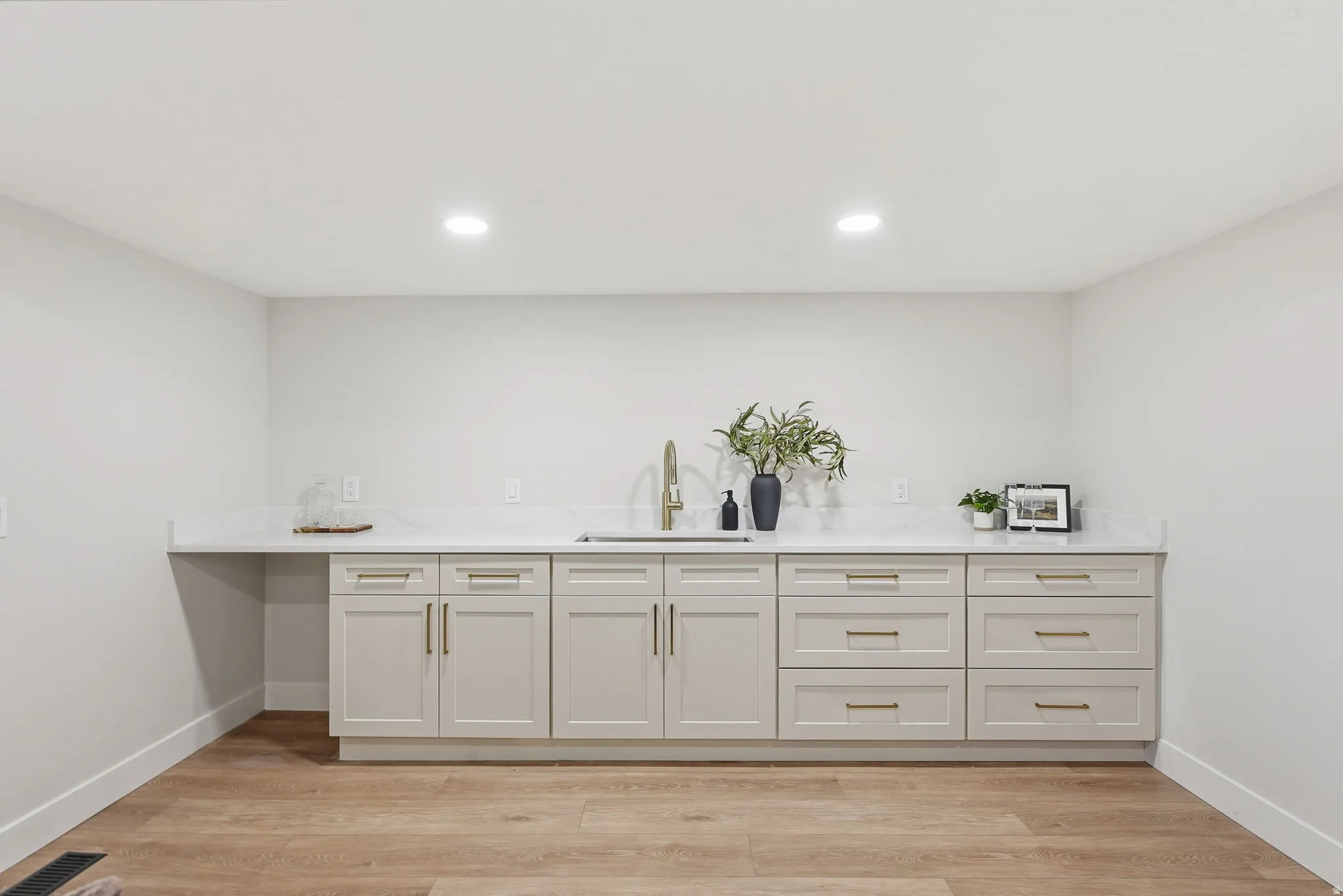 Contemporary wet bar featuring a single basin stainless steel sink with a gold-tone faucet, light gray cabinetry with gold-tone hardware, and a light-toned countertop