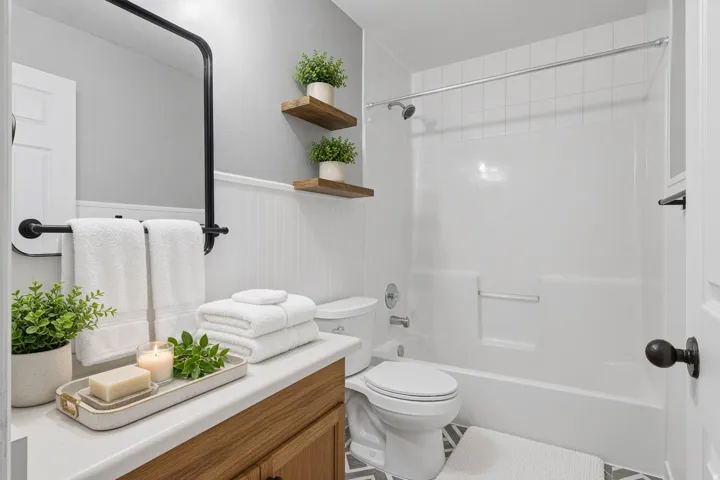 Built-in bathtub with shower, wood-finish vanity with white countertop, black-framed rectangular mirror, decorative wall paneling, and geometric pattern floor tiles