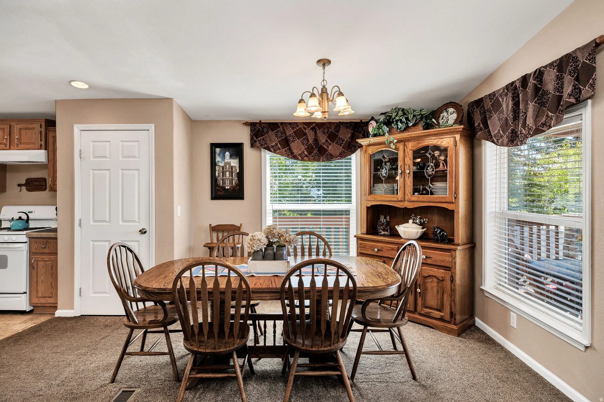Carpeted dining area featuring a chandelier, two windows with blinds, and a built-in hutch with glass-fronted upper cabinets