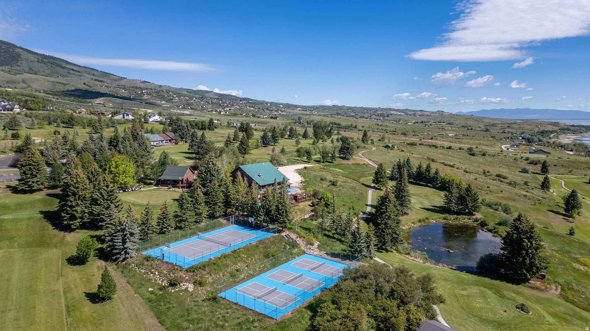 Aerial perspective revealing multiple tennis courts with blue surfaces, a green-roofed structure, and a circular pond