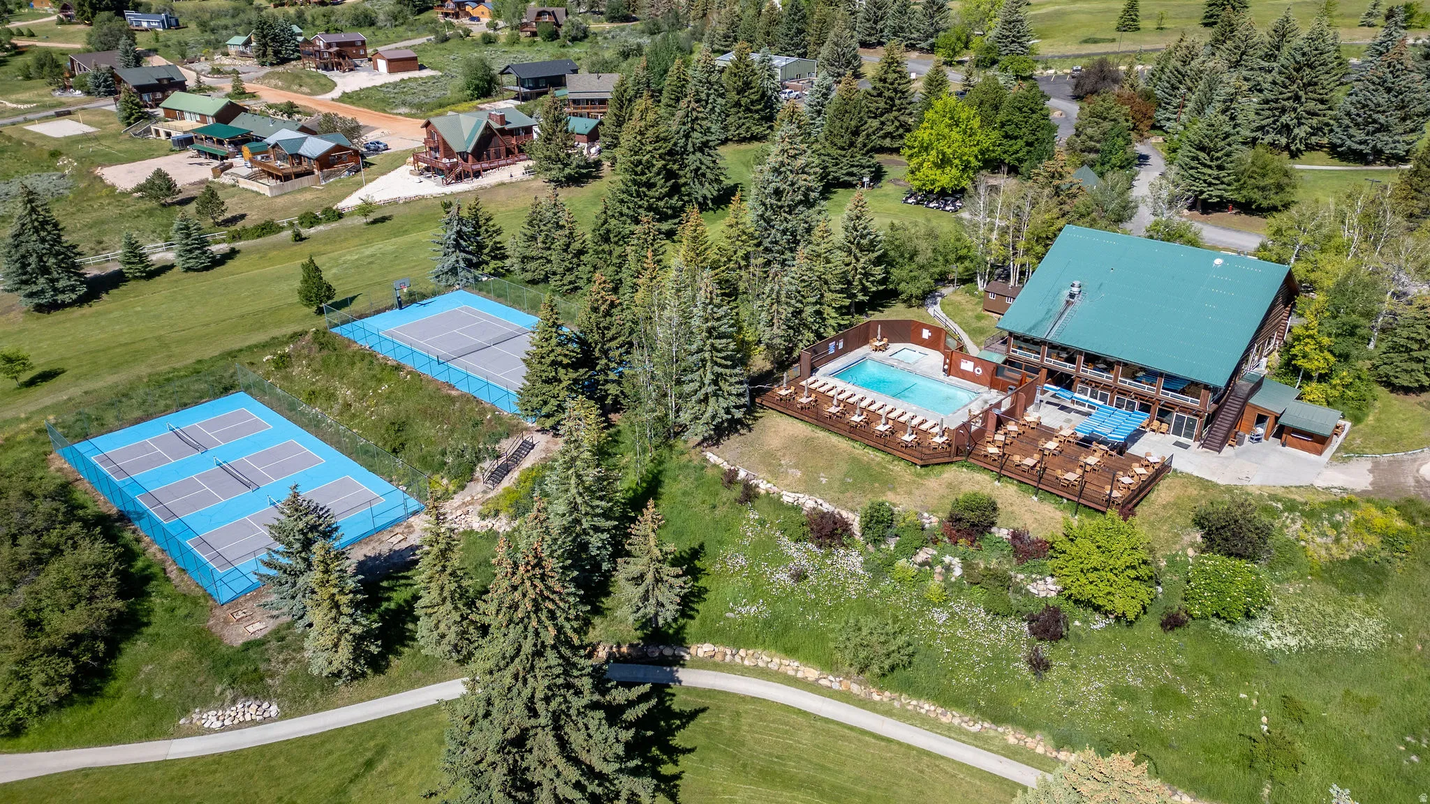 Expansive property featuring two blue-surfaced tennis courts, a large swimming pool with surrounding deck, and a multi-level building with a green metal roof