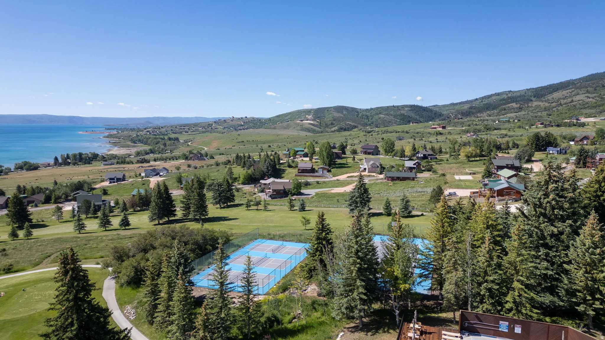 Expansive mountain landscape featuring a large body of water, distant residential properties, mature evergreen trees, and recreational courts