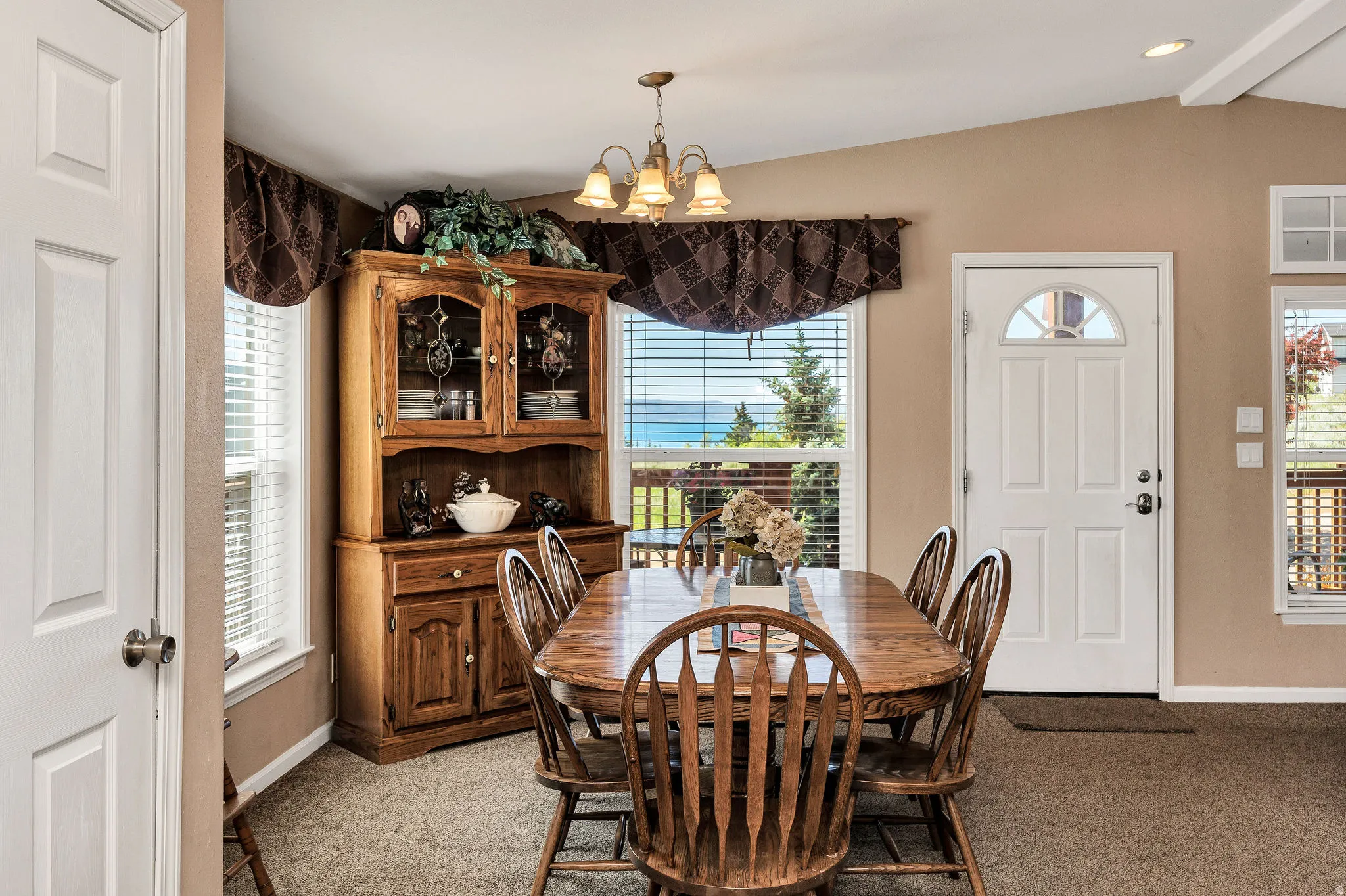 Spacious interior featuring high ceilings, light brown carpeting, multiple windows with horizontal blinds, and a white paneled exterior door with an arched glass insert