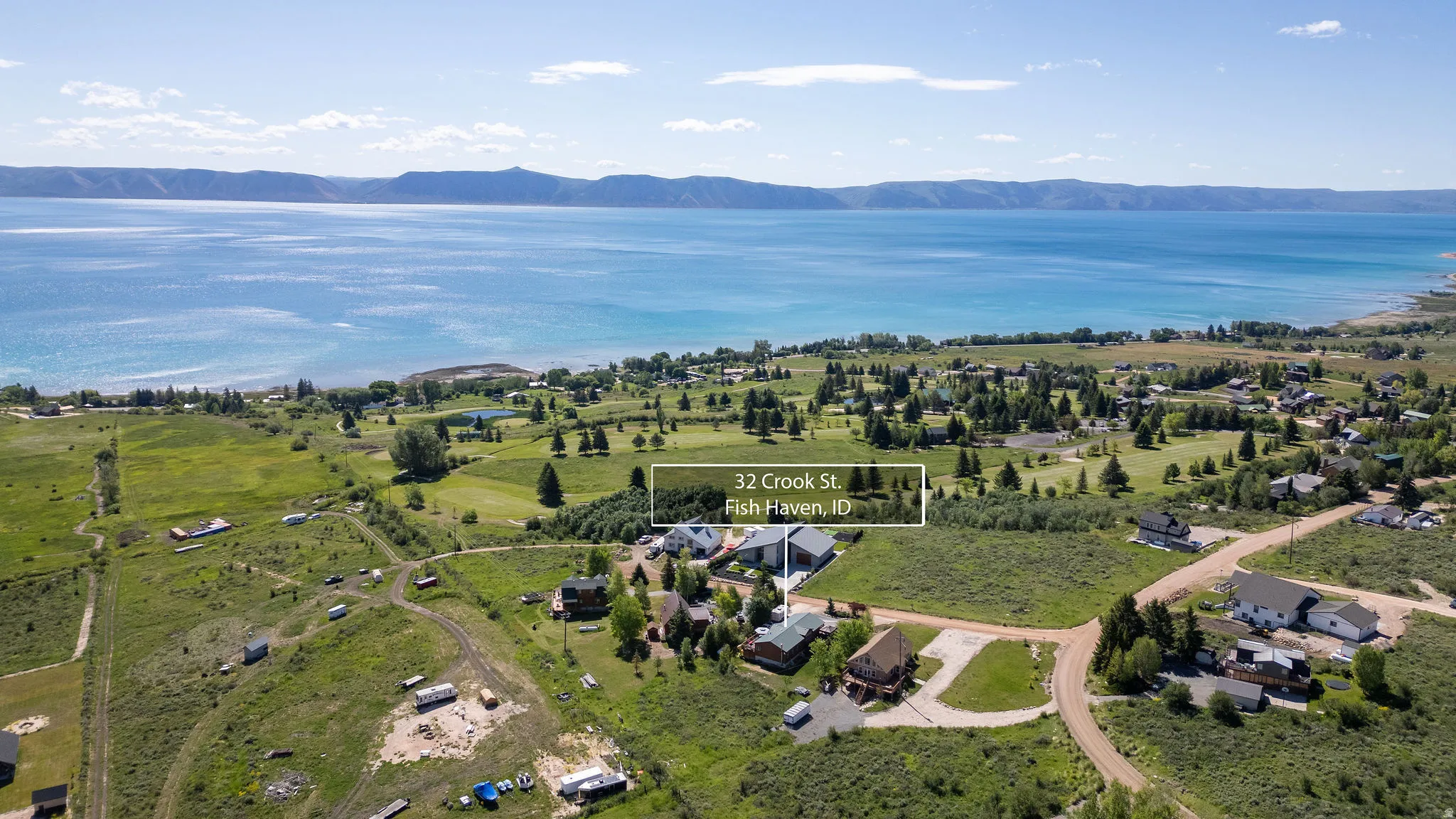 Aerial perspective showcasing a property in a waterside community, featuring a large body of water, expansive green spaces, surrounding mountainous terrain, and clustered residential structures
