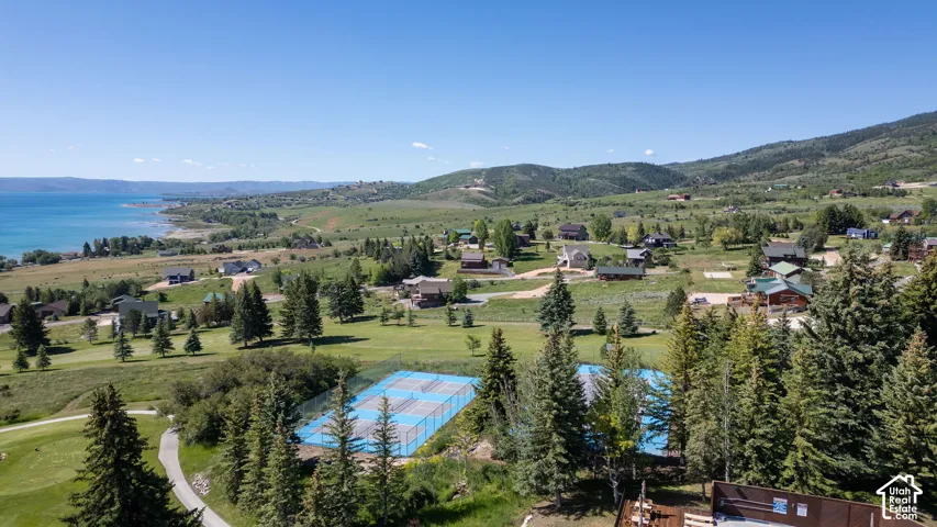 Expansive mountain landscape featuring a large body of water, distant residential properties, mature evergreen trees, and recreational courts
