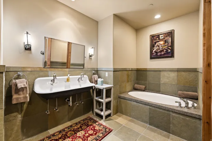Bathroom featuring a wall-mounted trough sink with two faucets, a built-in soaking tub with a stone-finish deck, slate-style wall tiling, and coordinating floor tiles